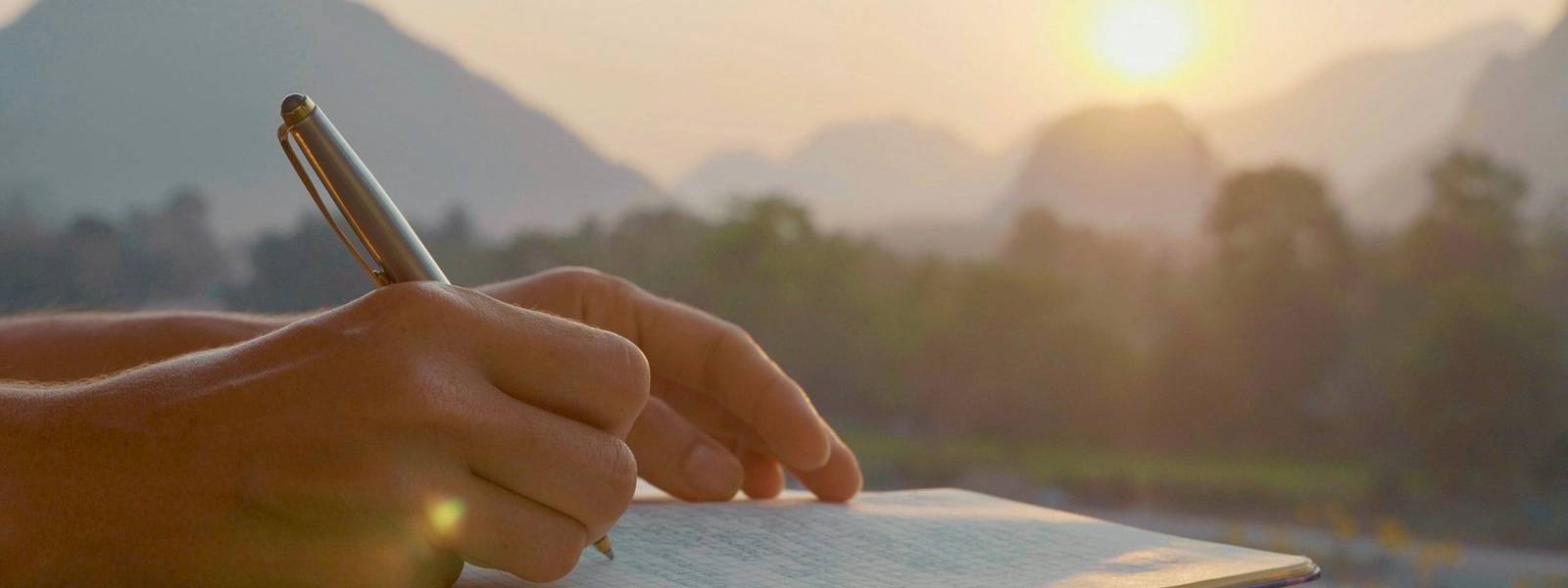Young woman writing travel notes in diary during sunrise with beautiful sun light and mountain landscape on the background, …