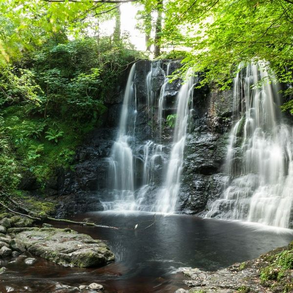 Waterfall with small water pond below surrounded by trees and lush vegetation in a deciduous forest in summer. Glenariff For…