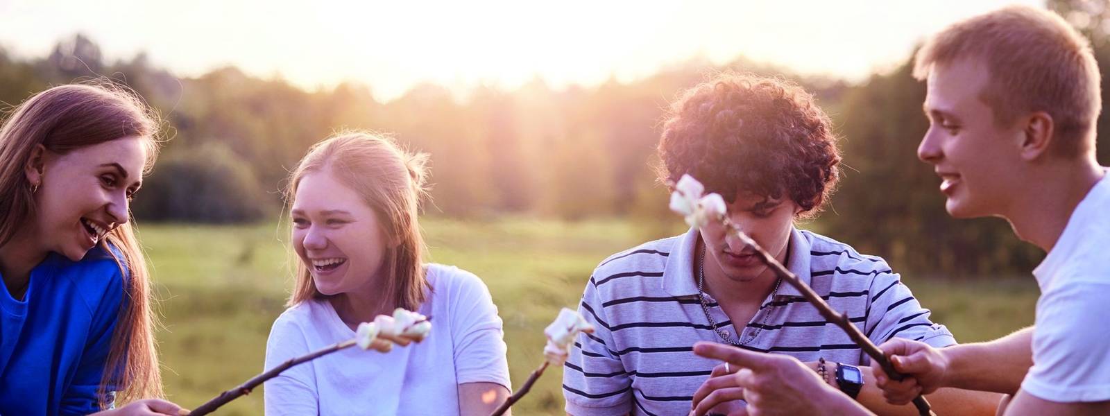 Photo of friendly company of friends have picnic outdoor, roast marshmallows over campfire, have positive expressions, pleas…