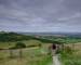 Cloudy overcast summer sunrise over the South Downs Way footpath from A middle aged male walker with backpack admiring the v…