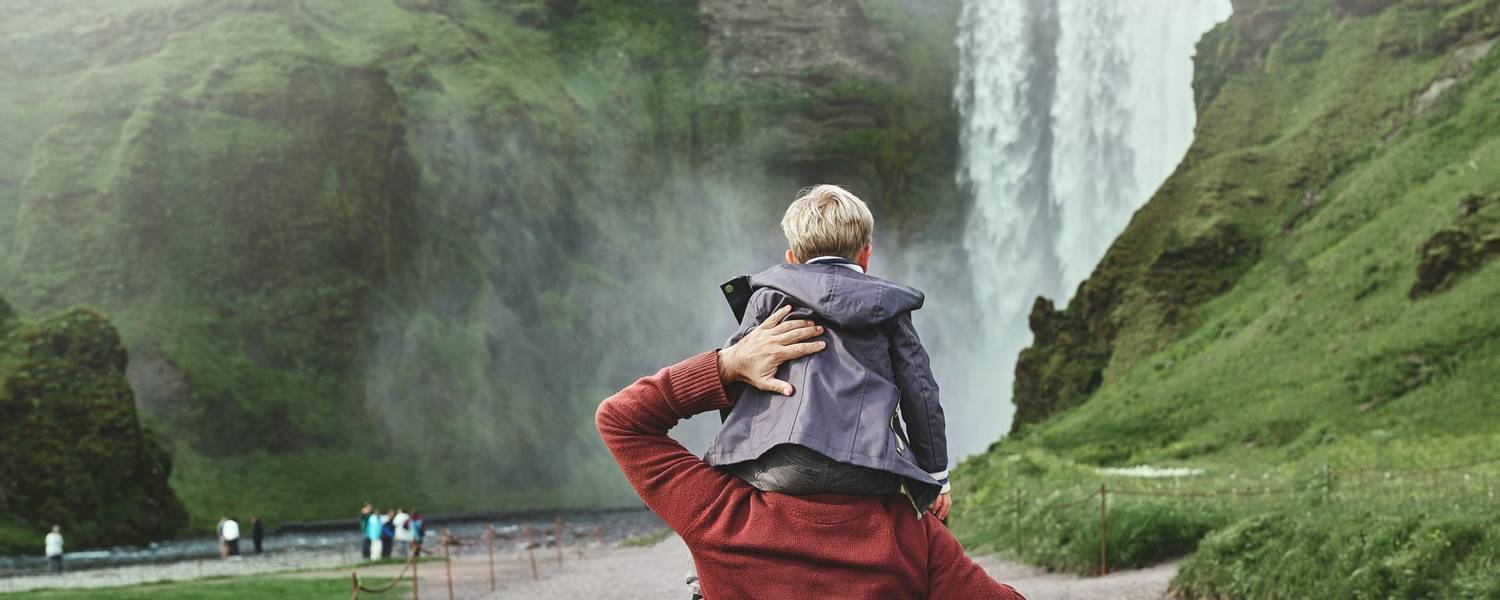 family in vacation traveling in Iceland. father carries a small son on his shoulders to the famous waterfalls of Skogafoss