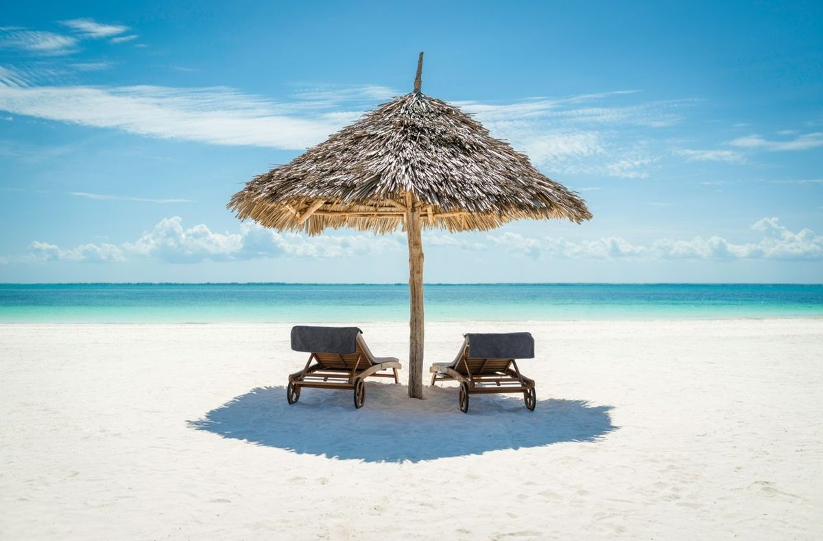 2 wooden sun loungers facing the tropical, turquoise blue Indian Ocean under a thatched umbrella on a white sandy Zanzibar b…