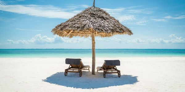 2 wooden sun loungers facing the tropical, turquoise blue Indian Ocean under a thatched umbrella on a white sandy Zanzibar b…