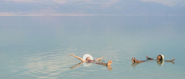 Lovely multiracial girls wearing sun hats, lying on back with outstretched arms, floating in salty water of Dead Sea. Unusua…