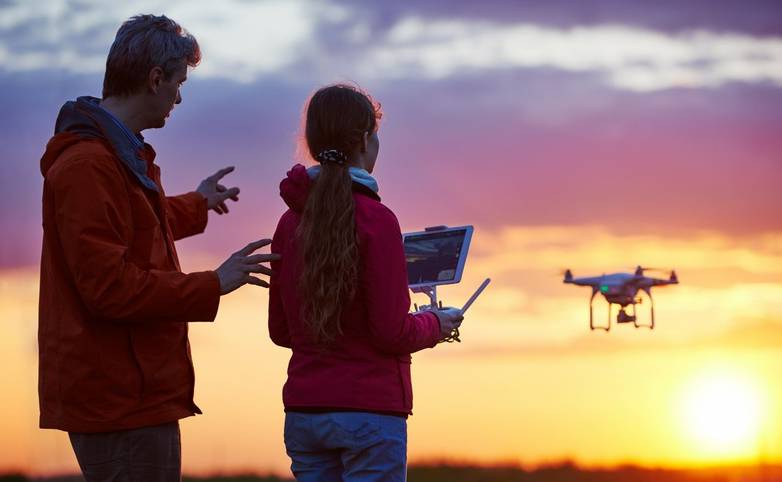 Man with child operating drone flying or hovering by remote control in sunset.
