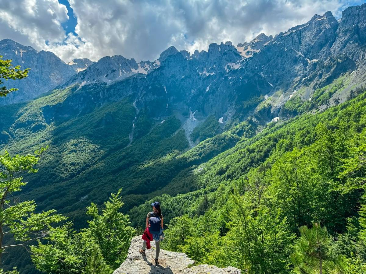 Hiker woman stands on edge of rock formation with panoramic view of Valbona Valley National park surrounded by majestic stee…