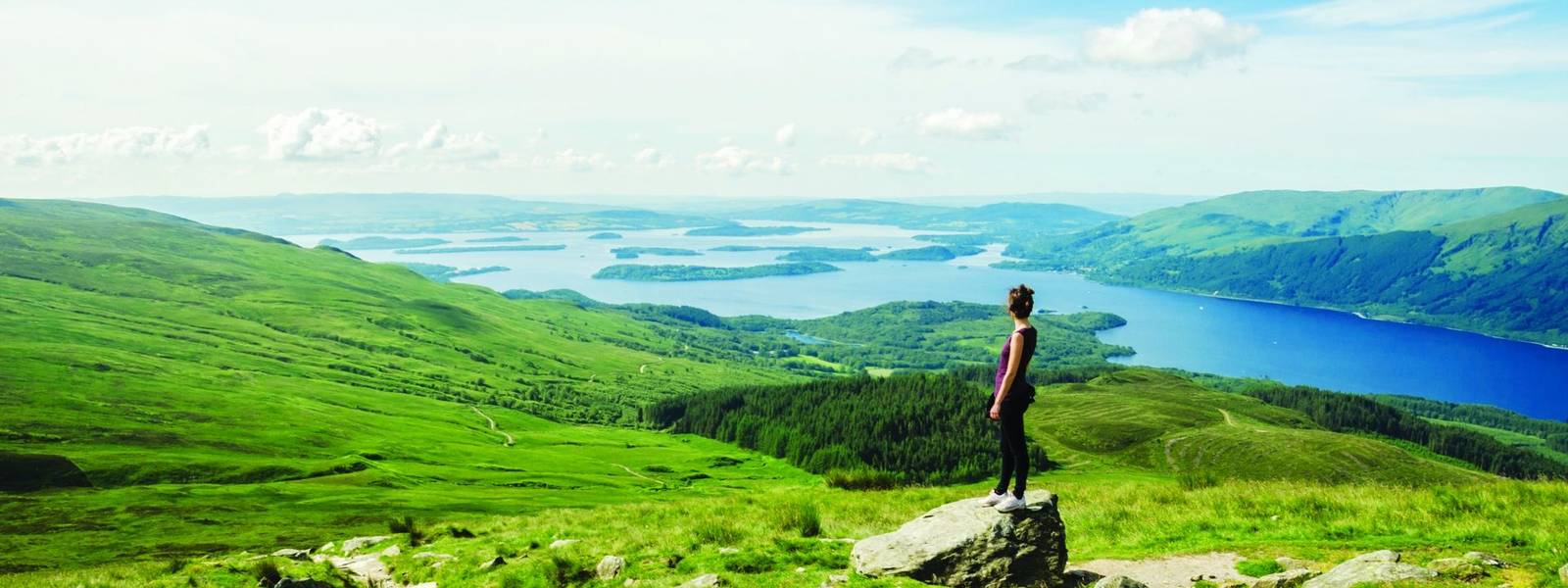 Female hiker admiring the landscape on a path leading to the top of Ben Lomond in a sunny day. Loch Lomond in the backgroun…