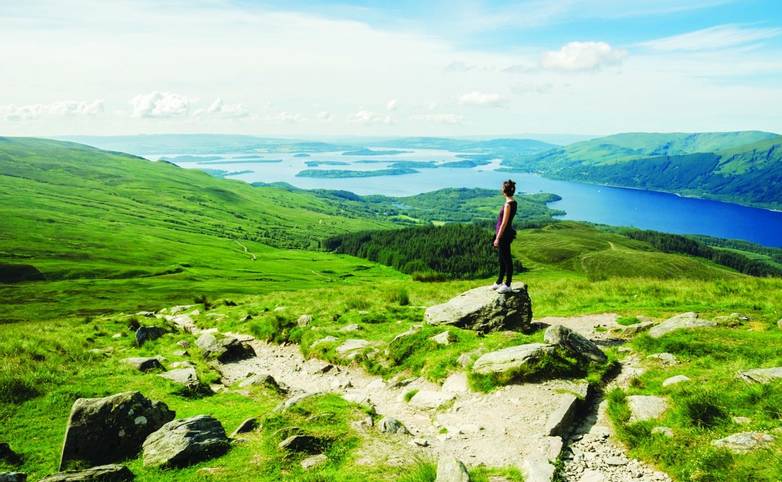 Female hiker admiring the landscape on a path leading to the top of Ben Lomond in a sunny  day. Loch Lomond in the backgroun…
