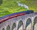 Close shot of Jacobite train travelling over the Glenfinnan Viaduct with a green hillsdie behind it and steam coming out of …