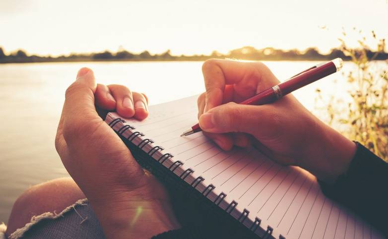 Close up hand of young woman with pen writing on notebook at riverside in the evening.