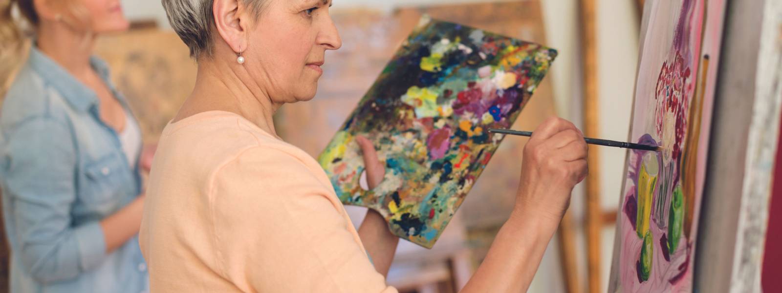 creativity, education and people concept - senior woman with brush and palette painting still life picture on easel at art s…