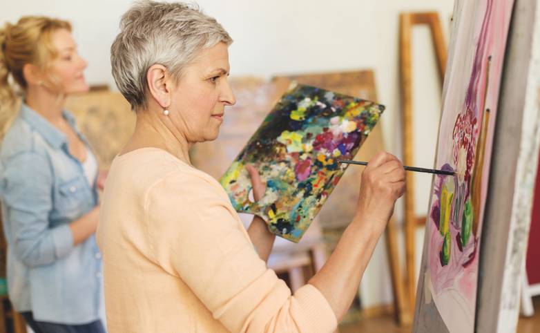 creativity, education and people concept - senior woman with brush and palette painting still life picture on easel at art s…