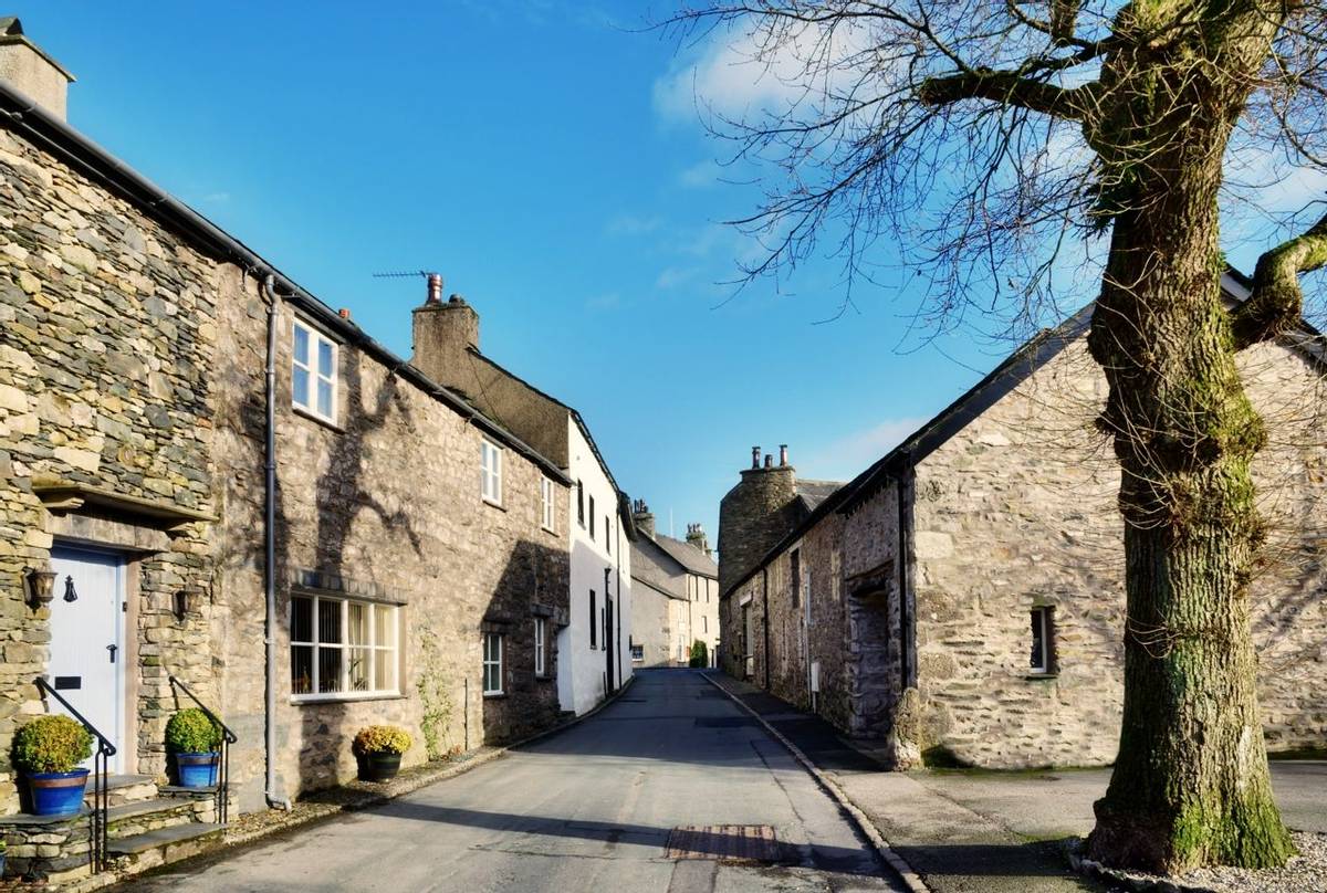 View of a picturesque street with cottages, and a tree set against a blue sky, in the village of Cartmel, Cumbria, England, …