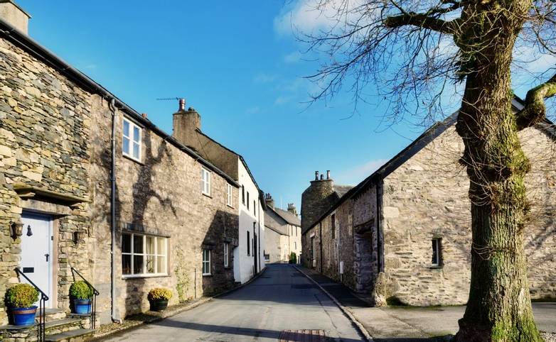 View of a picturesque street with cottages, and a tree set against a blue sky, in the village of Cartmel, Cumbria, England, …