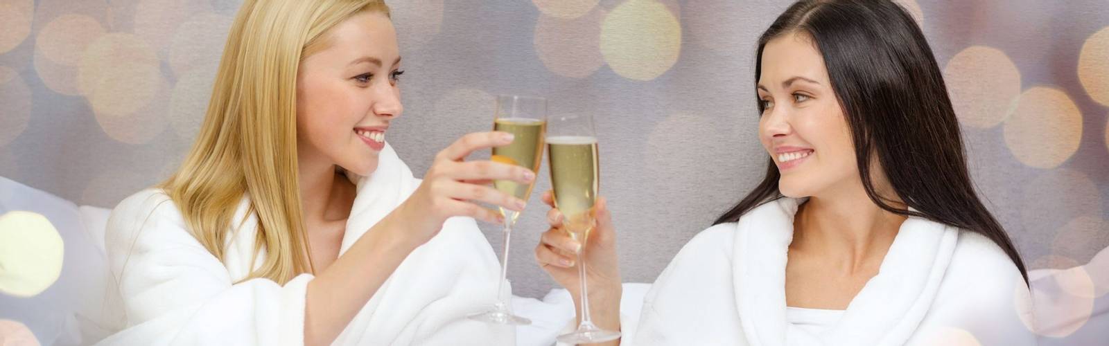 travel, celebration, friendship and holidays concept - smiling girlfriends in bathrobes with champagne glasses in bed at hot…