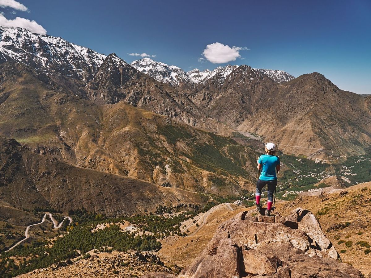 Caucasian blonde tourist girl on a hike looking towards snow covered High Atlas monuntain peaks with Jebel Toubkal in the mi…