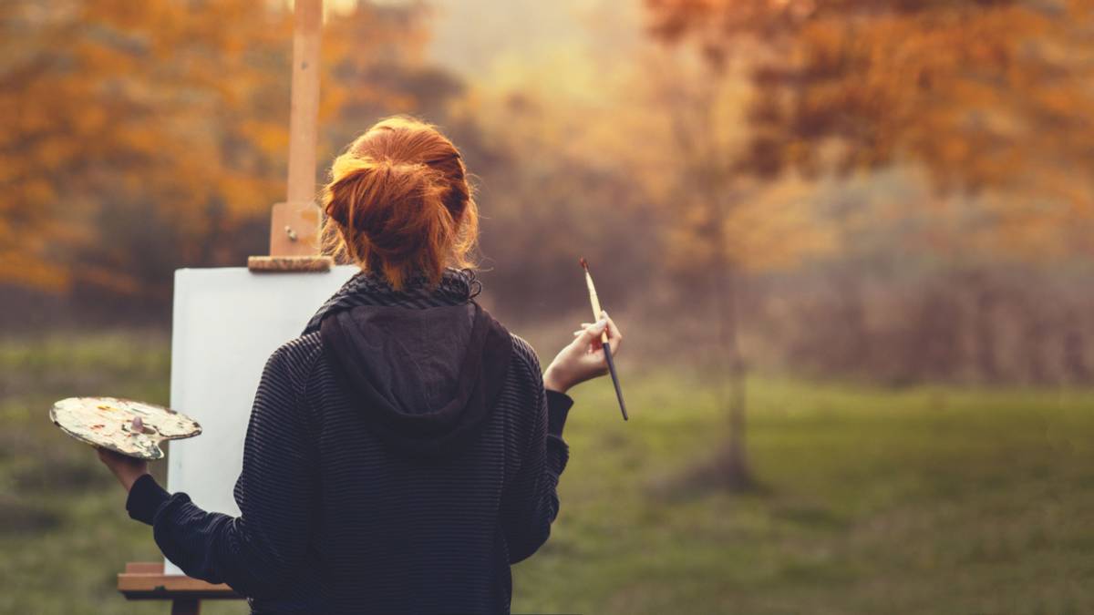 redhead girl drawing a picture on an easel in nature, young woman with a paint brush and a palette among autumn trees, a con…
