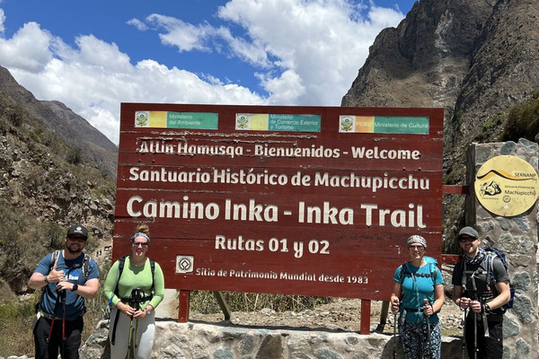 The Beginner's Guide to the Inca Trail Hike. Four people stood in front of a brown sign saying Camino Inka. Inka Trail. They are in the Andes with a backdrop of mountains.