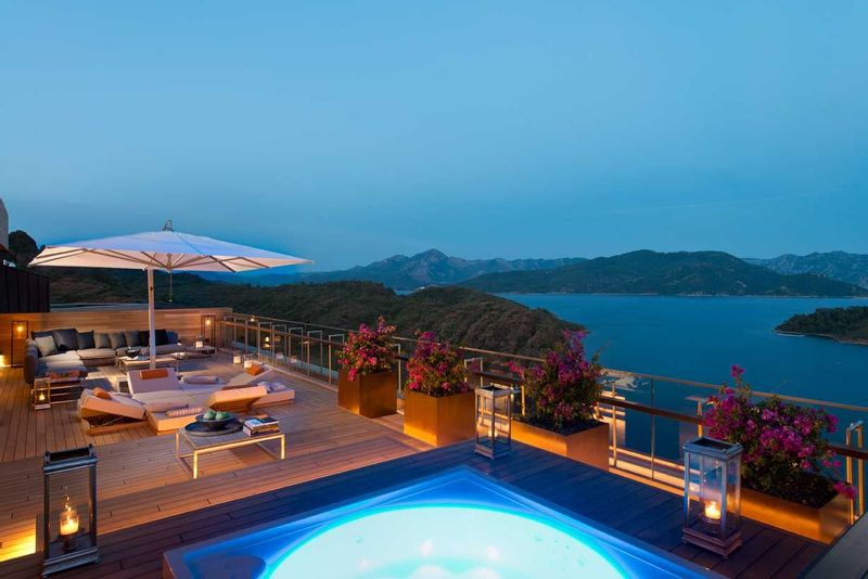 Landscape image of a pool and luxurious seating area overlooking a lake and mountains at nightfall